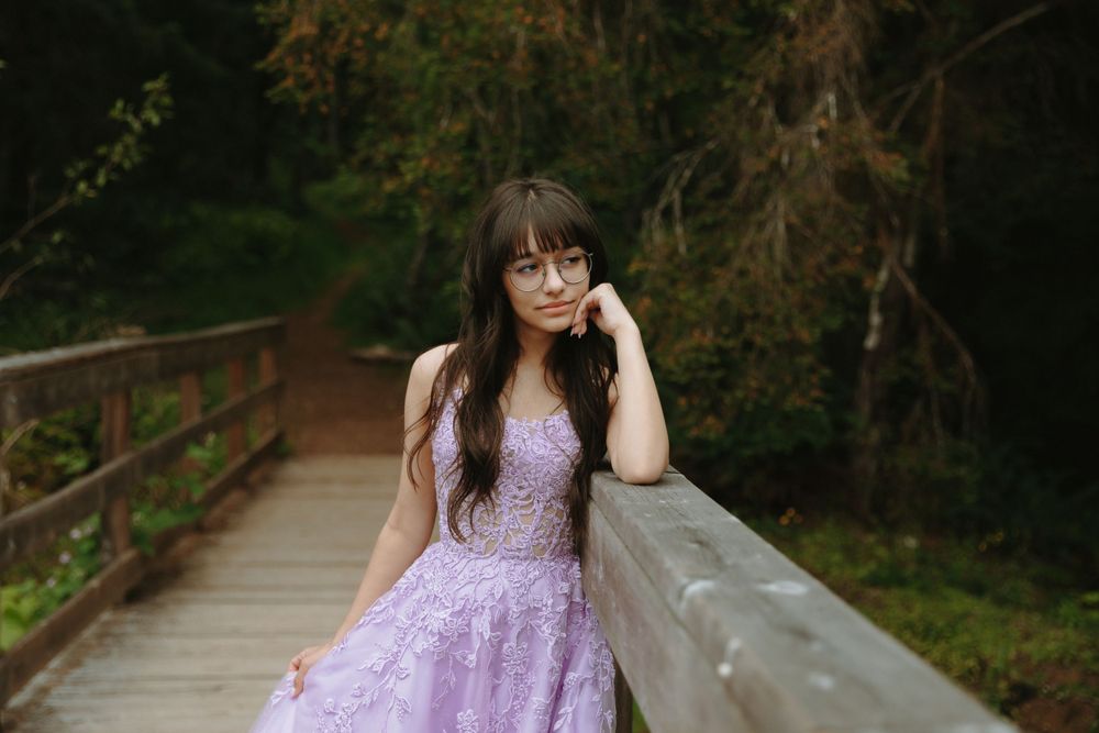 A person in a lilac lace dress leans against a wooden bridge railing surrounded by lush green foliage.