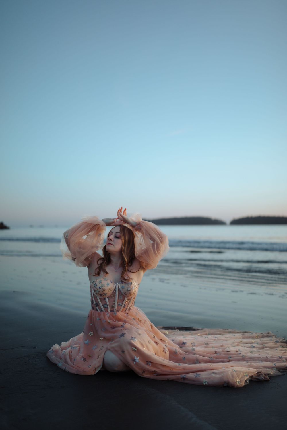 A dreamy beach portrait with a person in a flowing peach gown posing on dark sand at sunset.