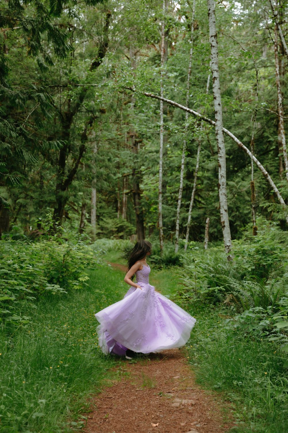 A flowing lavender ballgown twirls on a forest path surrounded by lush green trees and foliage.