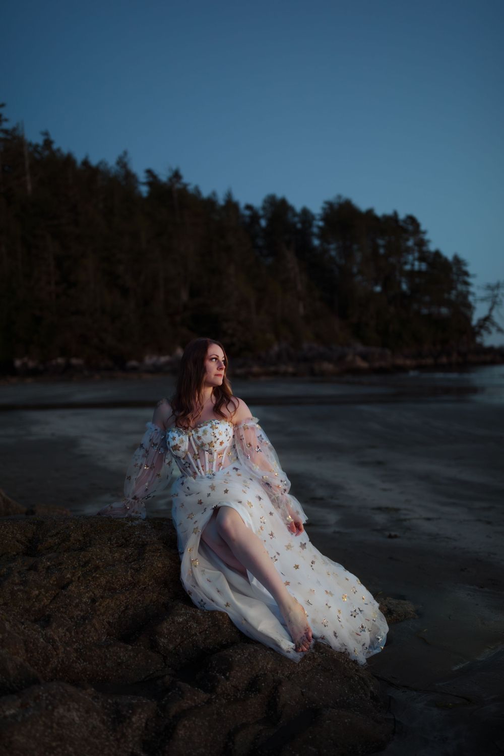 A dreamy fashion portrait on a rocky beach at dusk featuring a white floral dress with flowing sleeves.