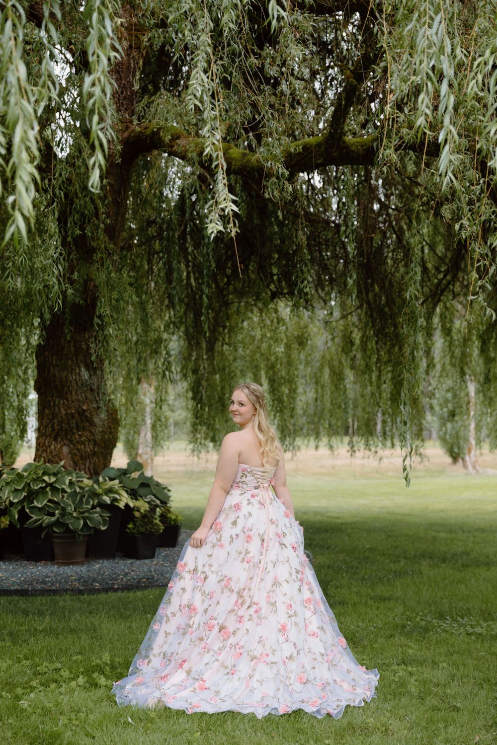 A floral ball gown with pink details is showcased under a graceful weeping willow tree on a manicured lawn.