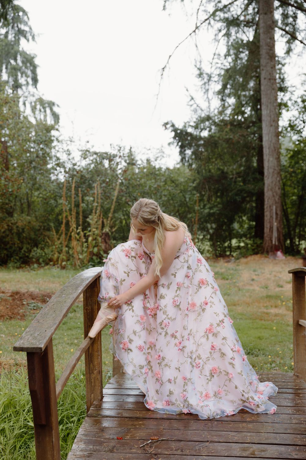 A person in a flowing floral dress bends down on a wooden bridge in a serene garden setting.