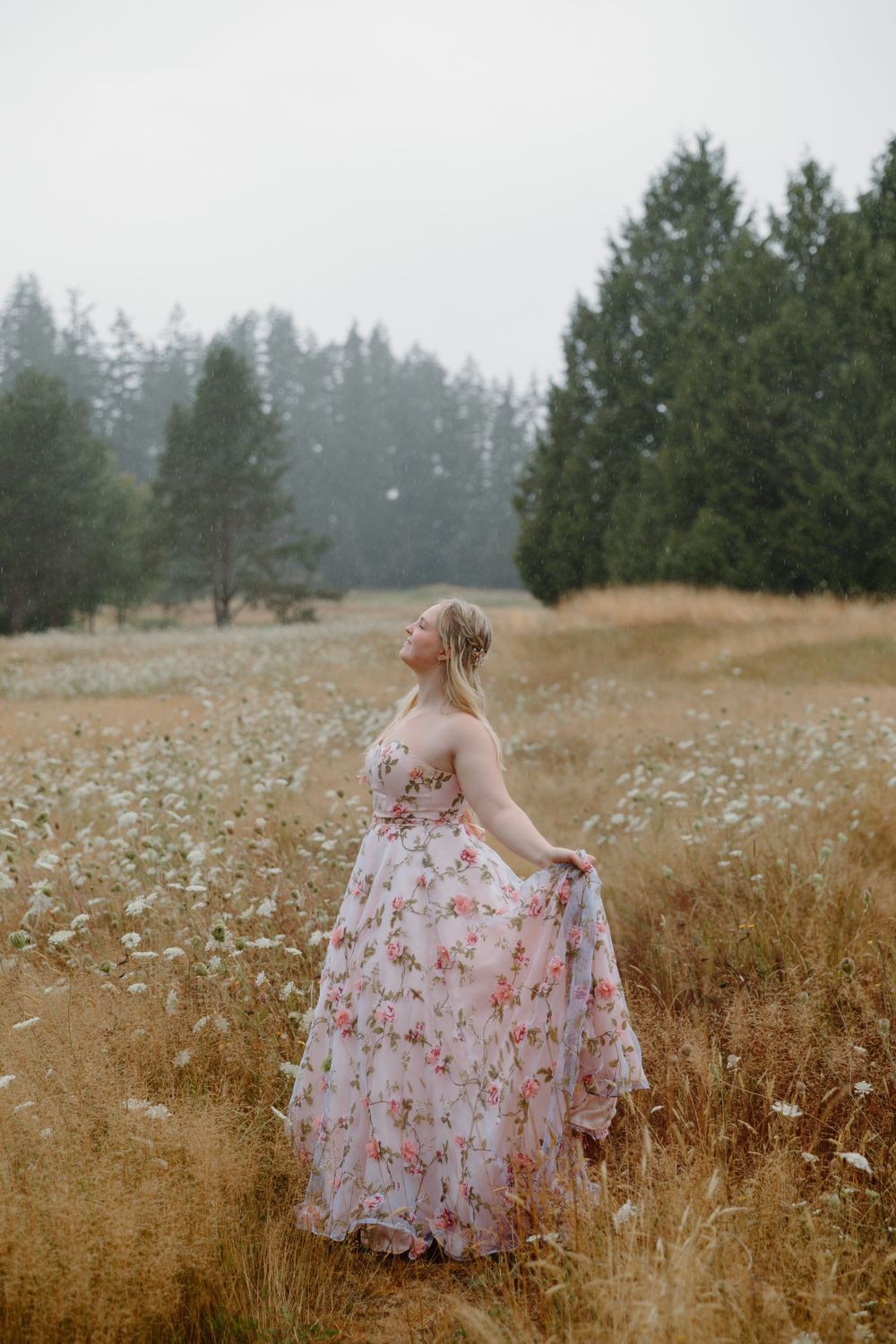 A person in a floral pink dress twirls in a misty meadow surrounded by evergreen trees.