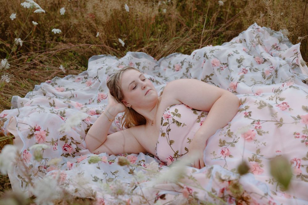 A figure rests outdoors on a floral-patterned pink blanket surrounded by dried grass and wildflowers.