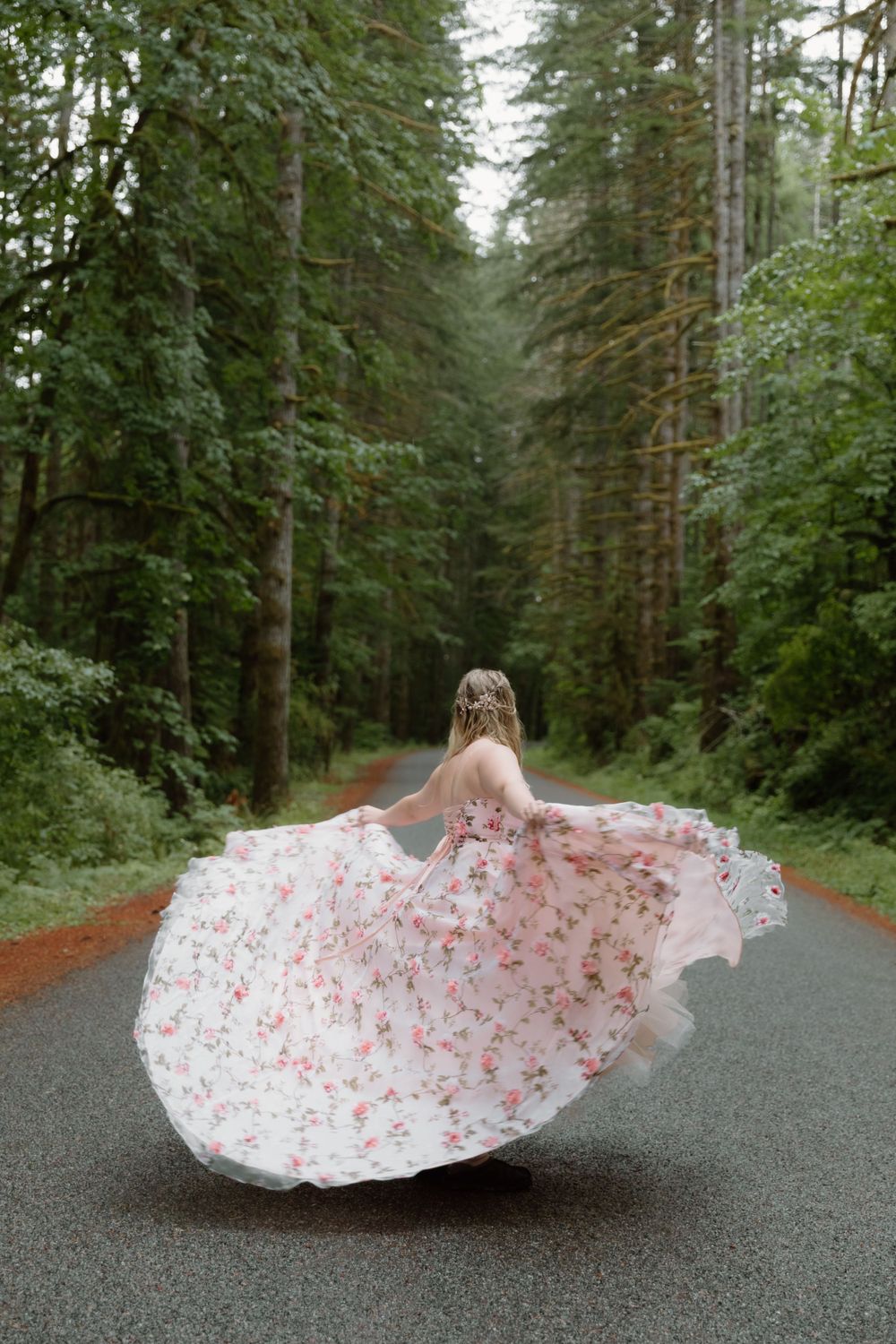 A floral white and pink dress twirls on a misty forest road surrounded by tall evergreen trees.