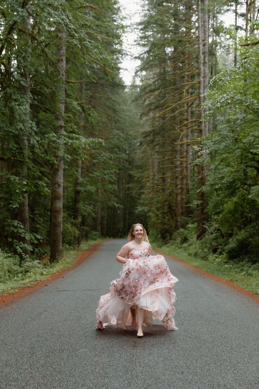 A person dances joyfully in a floral pink dress on a forest road lined with tall evergreen trees.