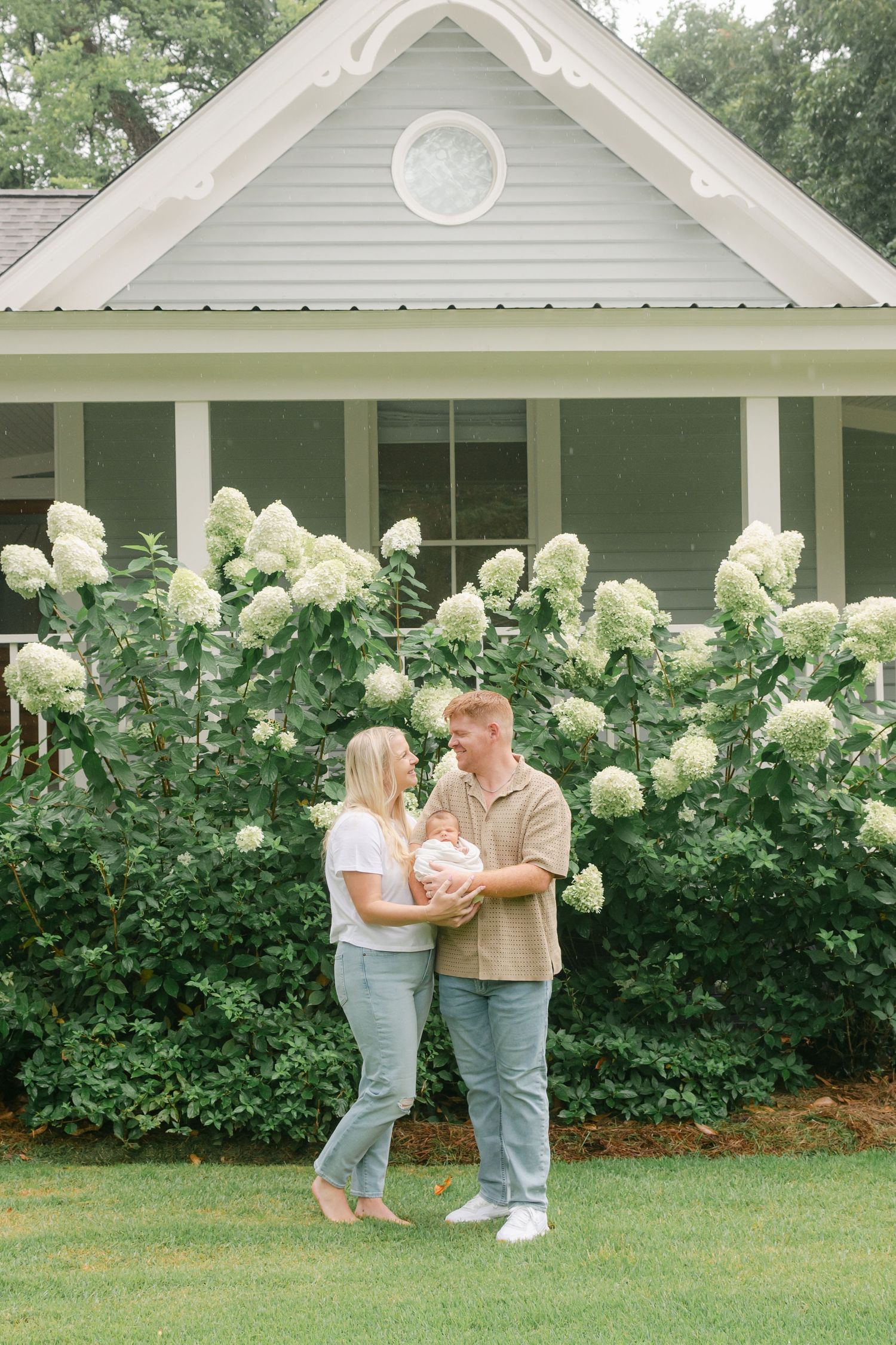 A young couple holds their newborn in front of blooming white hydrangeas outdoors.