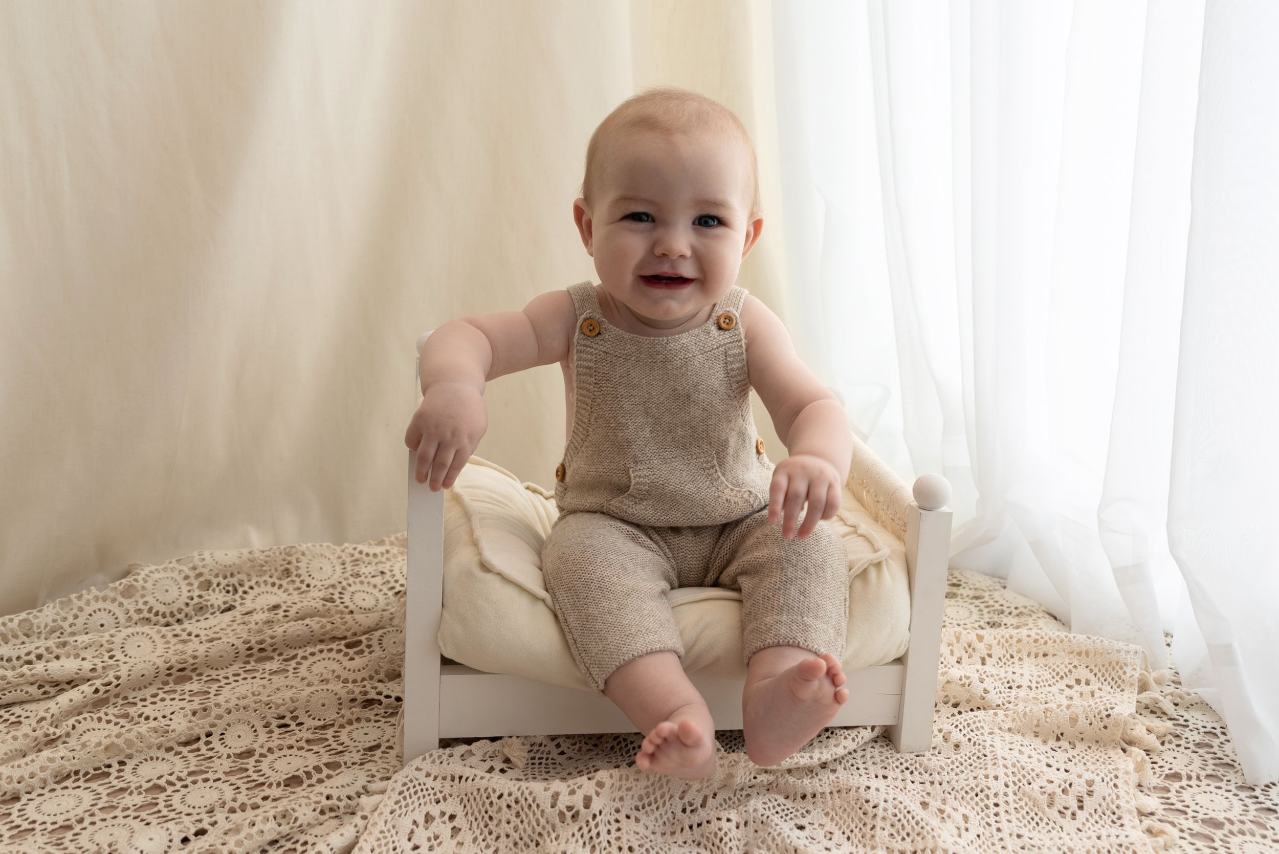 Smiling baby sitting on small white chair wearing beige outfit against neutral background and textured rug.