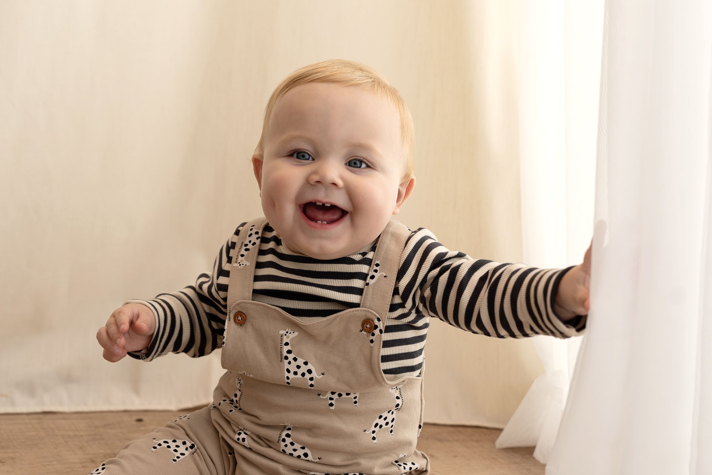 Baby wearing striped shirt and overalls laughs joyfully while sitting against white wall.