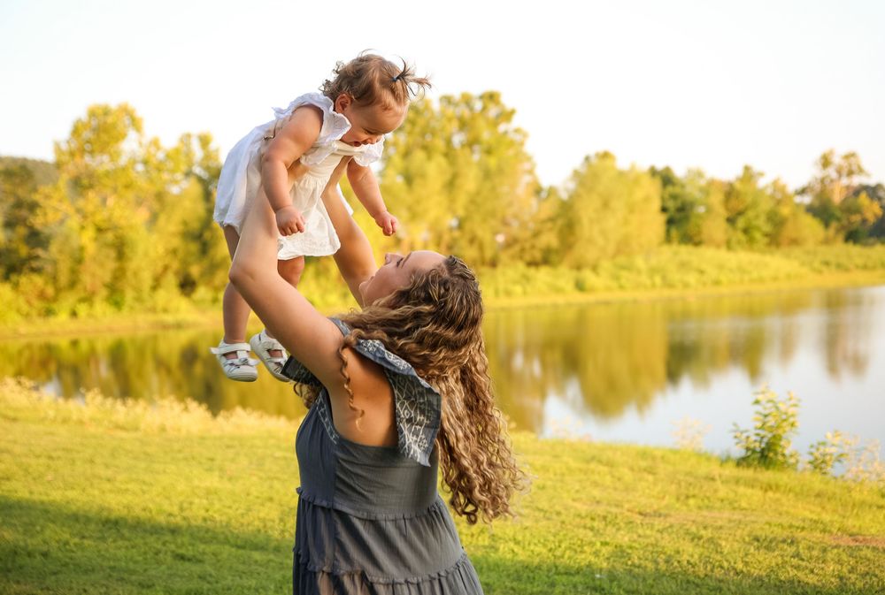 Mother joyfully lifting baby girl in white dress above her head near a sunlit lake.