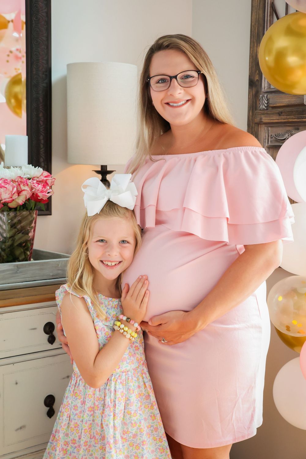 Smiling pregnant woman in pink dress posing with young blonde girl at a baby shower.
