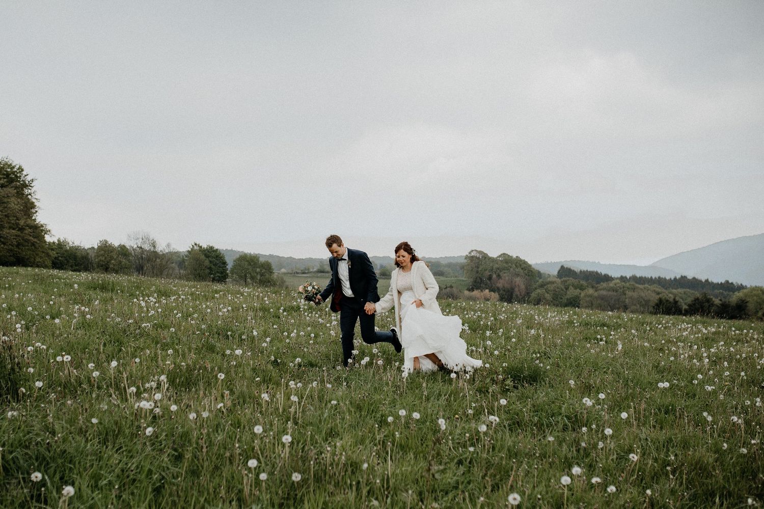 A couple walks hand in hand through a green mountain meadow filled with wildflowers on their wedding day.