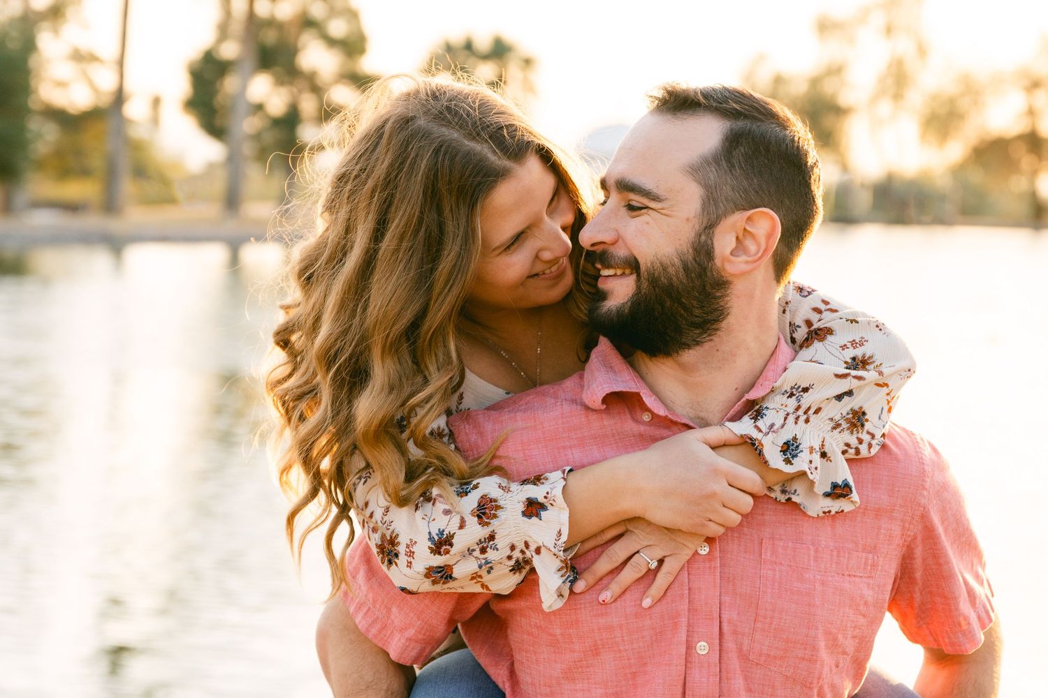 A man in a pink shirt is looking at a woman that he's carrying on his back. They're both smiling and there's a lake and palm trees nearby.