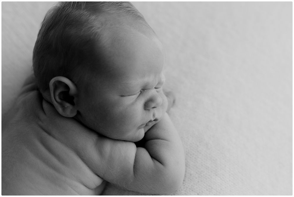 Black and white newborn photo of a sleeping baby with hands tucked under chin on soft blanket.