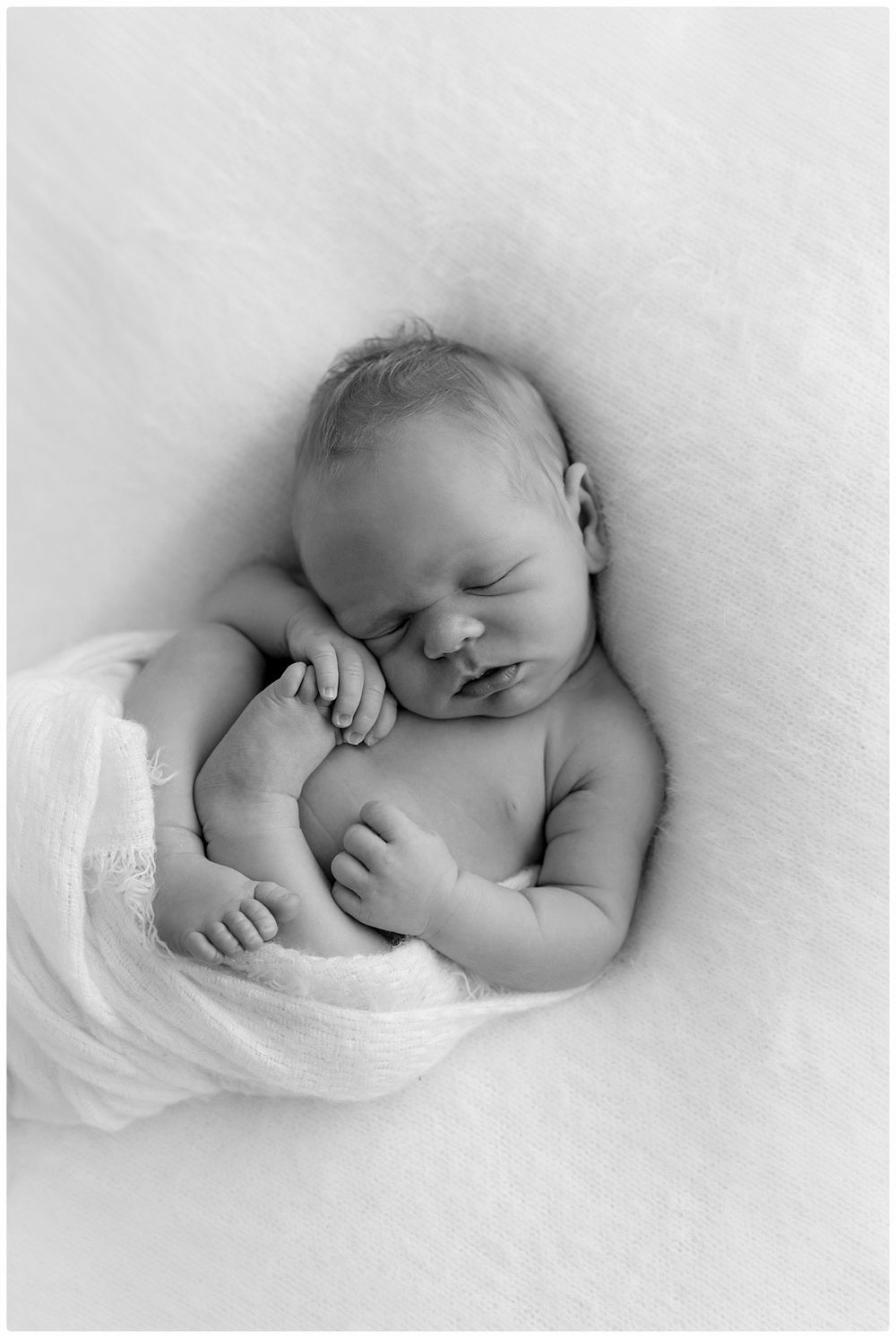 Black and white newborn photo of a sleeping baby wrapped in a soft white blanket on a fluffy white background.