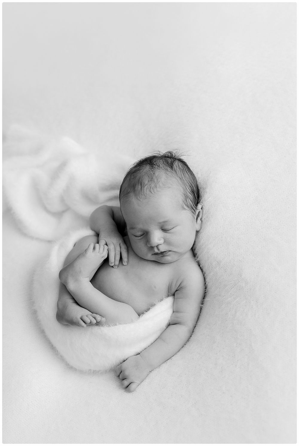 Black and white newborn photo of a sleeping baby curled up on a soft white fluffy blanket.
