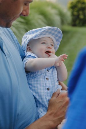 Baby in blue gingham outfit being held up against sunny outdoor background.