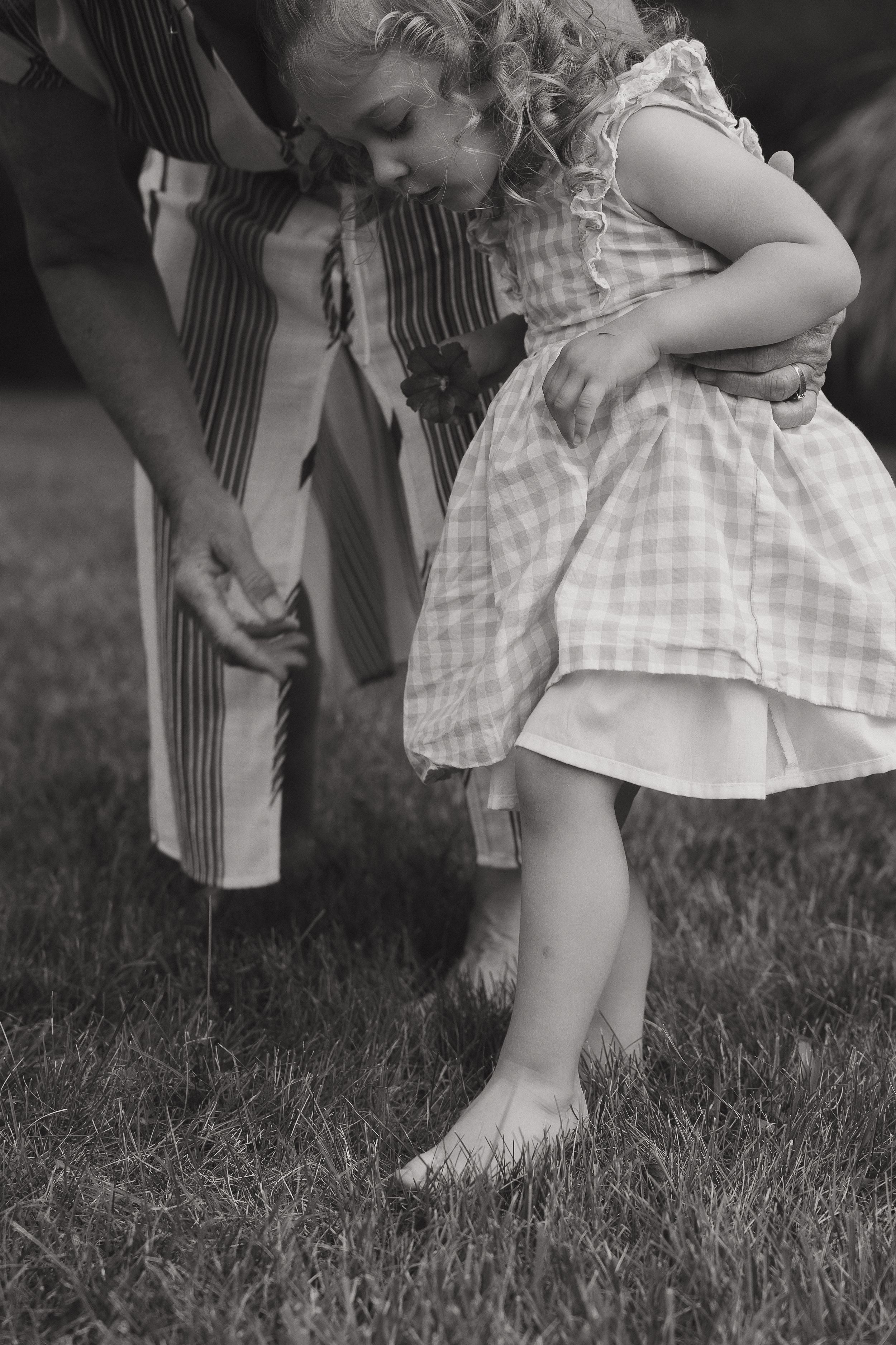 Black and white photo of small child in checkered dress bending down to touch grass.