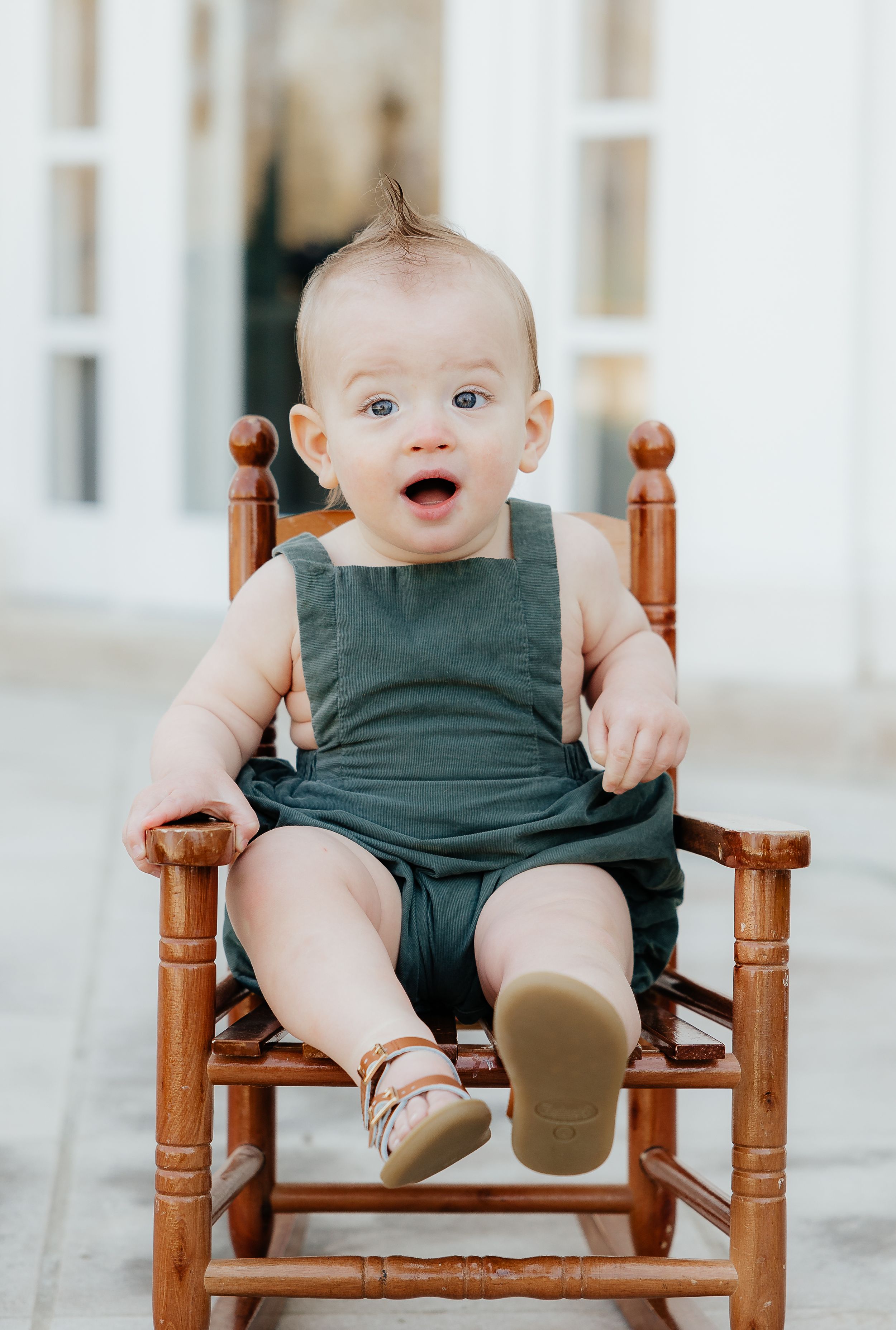 Baby wearing dark green romper sitting in wooden rocking chair.