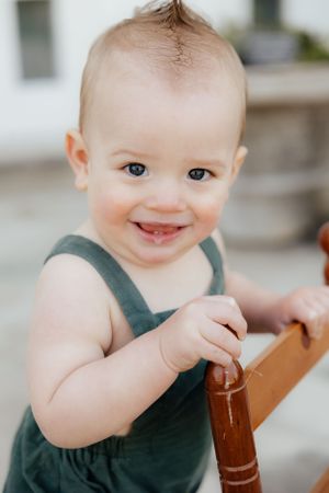 Baby in dark green outfit smiling while sitting in wooden chair.