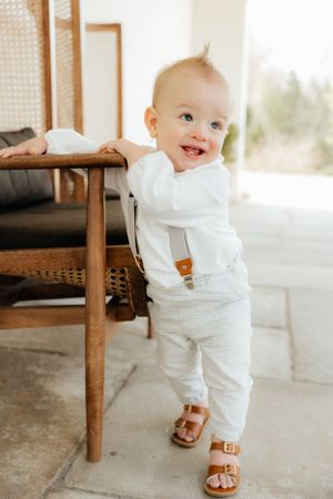 Baby in white outfit and sandals standing beside wooden chair in bright interior.