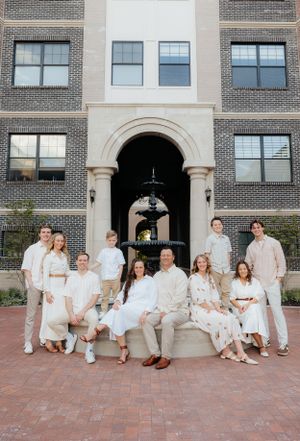Large family group in white clothing sits casually along brick building entrance.