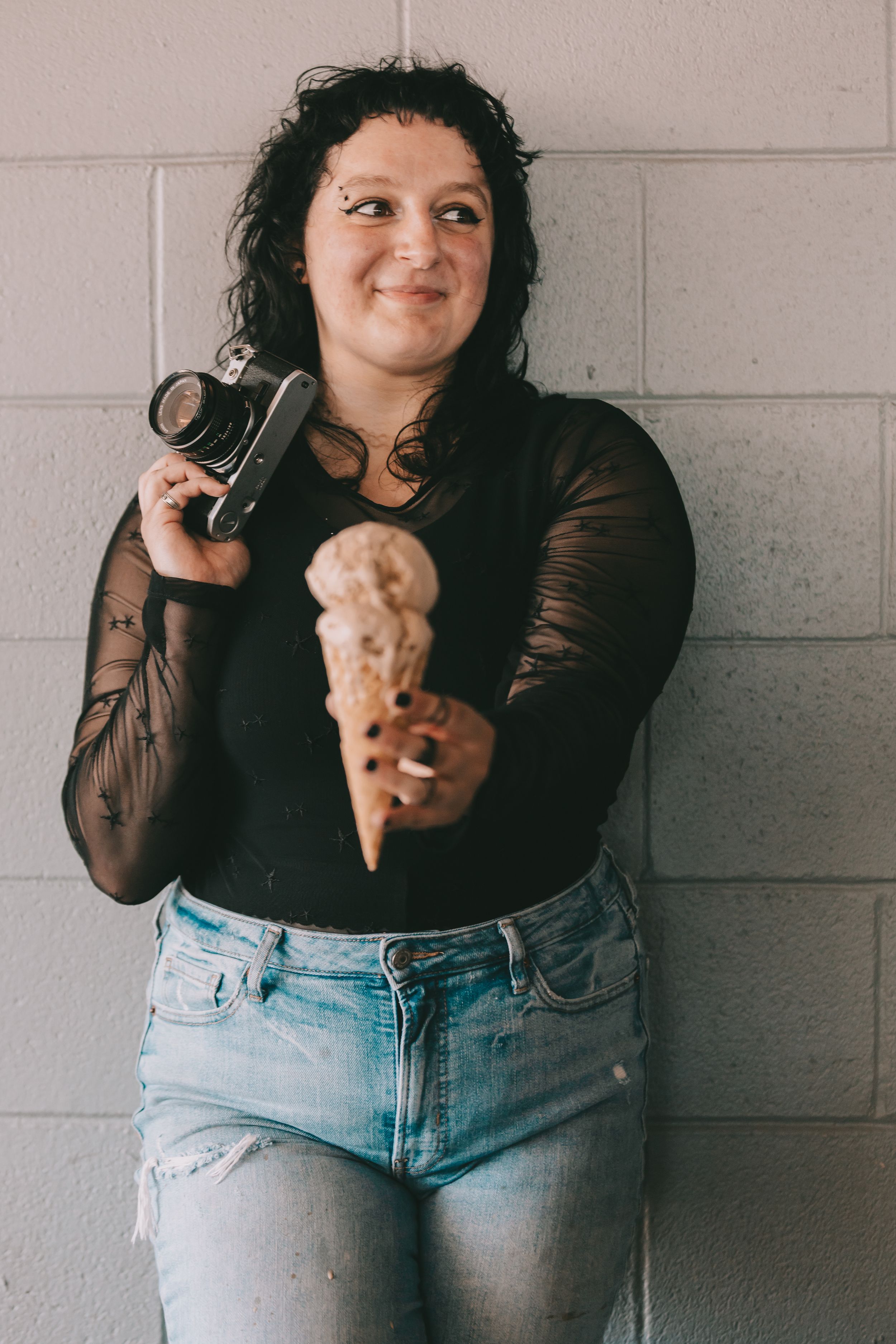 An image of a woman holding a camera and an ice cream cone standing in front of a grey brick wall.