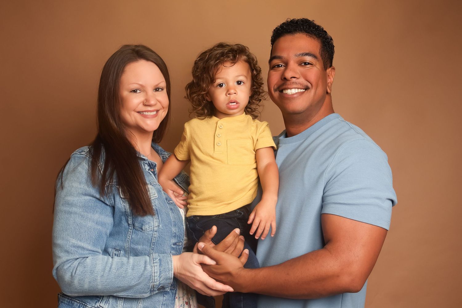 A diverse family in matching blue shirts poses together against a brown studio backdrop.