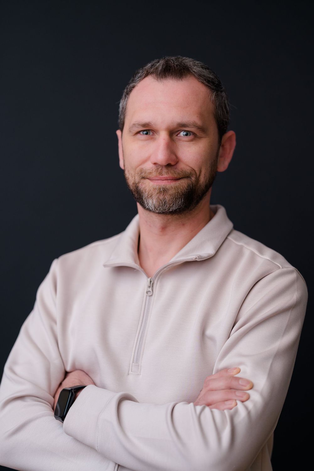 A middle-aged man with a beard in a beige zip-up top, arms crossed, smiling against a dark background.