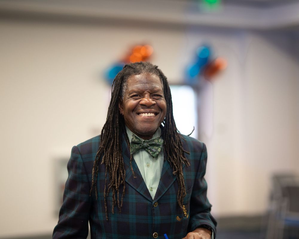 A person in a plaid jacket and bow tie smiles warmly at an indoor event.