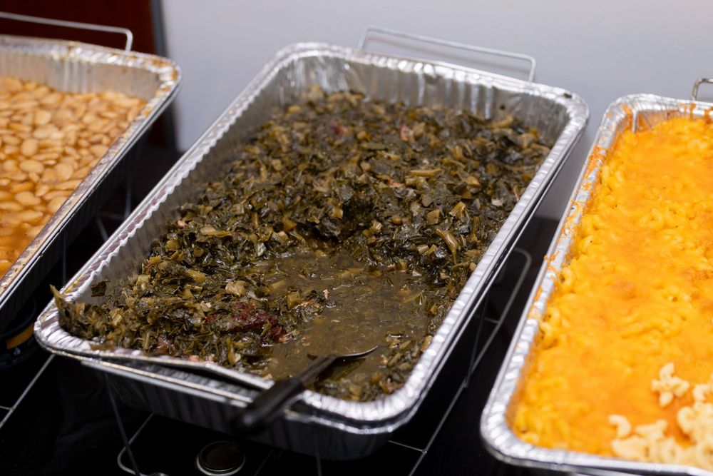 Three catering trays filled with macaroni and cheese, collard greens, and corn dishes.