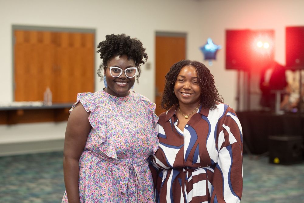 Two people in stylish dresses pose together at an indoor event with patterned fabric and decorative lighting.