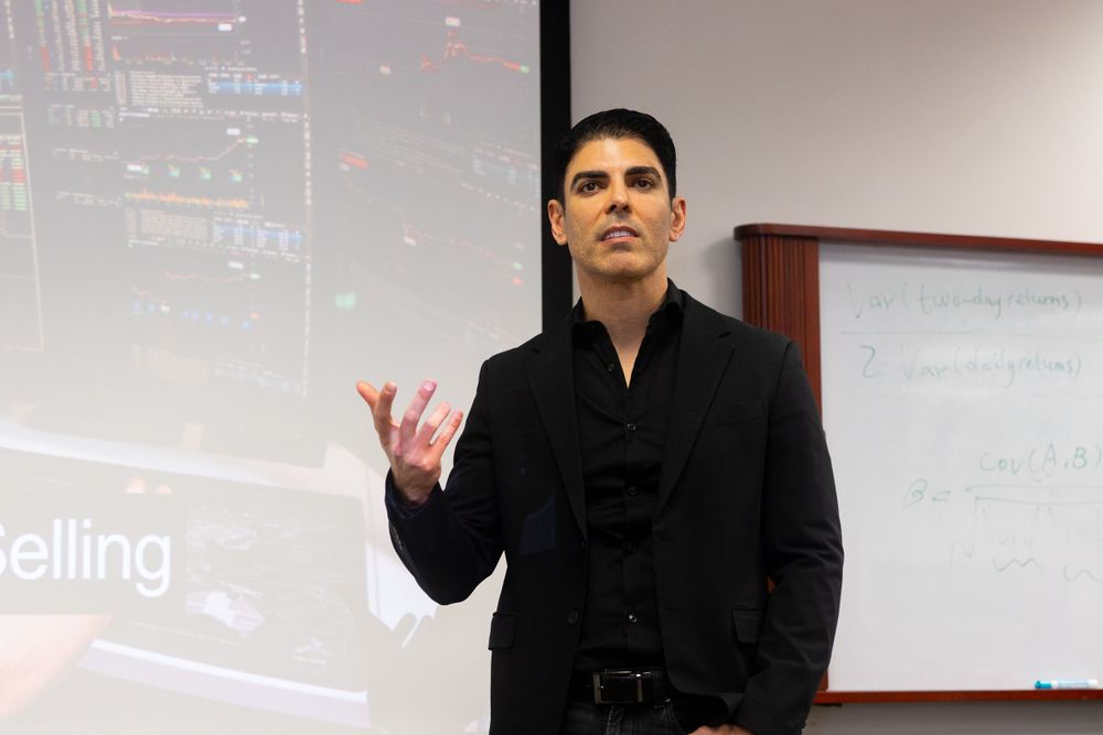 A speaker in a black outfit gestures while giving a presentation in front of a whiteboard in a classroom setting.