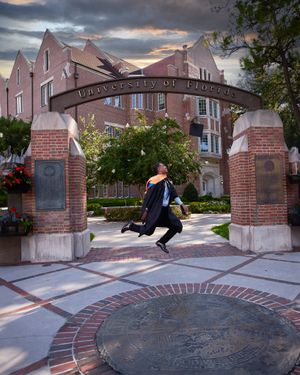 Graduate performs joyful jumping pose under university entrance arch at dusk.