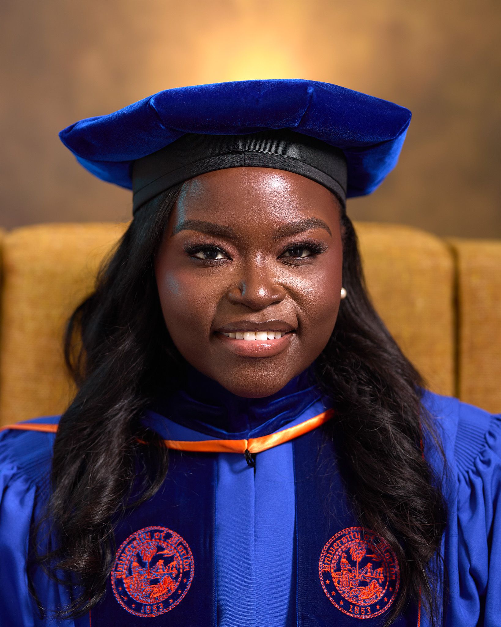 Graduation portrait in blue and orange academic regalia against warm background.