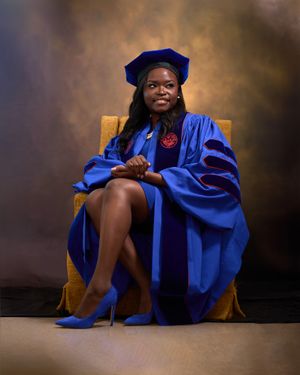 Graduate wearing blue doctoral regalia poses regally in chair against dark dramatic background.