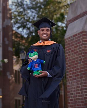 Graduate in academic regalia holds plush mascot toy while posing against brick wall.