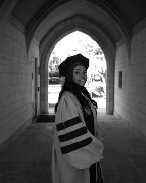 Graduate in white academic regalia stands confidently beneath arched stone passageway.