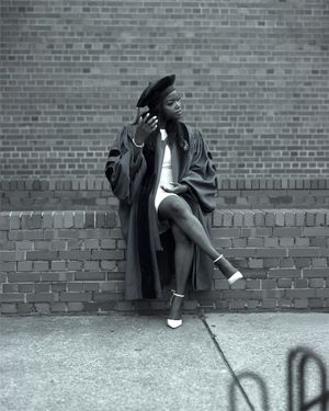 Graduate in black academic regalia poses stylishly against brick wall in monochrome photo.