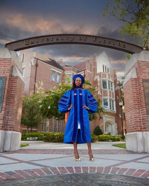 Graduate in blue academic regalia stands proudly under the University of Florida entrance archway.