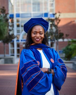 Graduate in blue doctoral regalia poses outdoors on campus with bright smile.