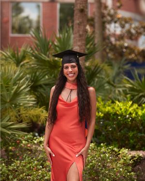 Graduate in coral dress and cap poses amid tropical landscaping.