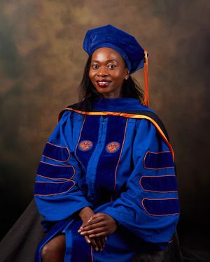 Graduate in blue doctoral regalia with orange trim poses against brown backdrop.
