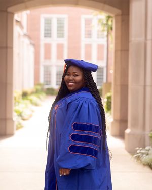 A graduate in royal blue doctoral regalia poses under an archway at a university.
