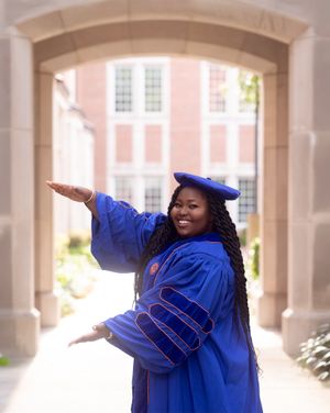 A joyful graduate in blue doctoral robes strikes a celebratory pose under an archway.
