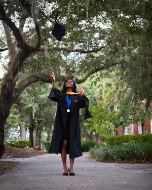 Graduate throws graduation cap in celebratory pose on tree-lined campus walkway.