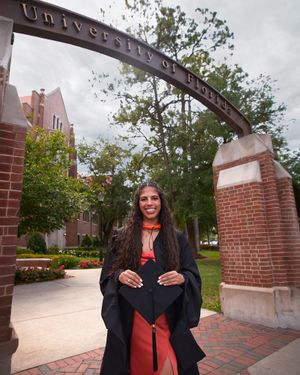 Graduate in coral dress poses beneath University of Florida arch during graduation.