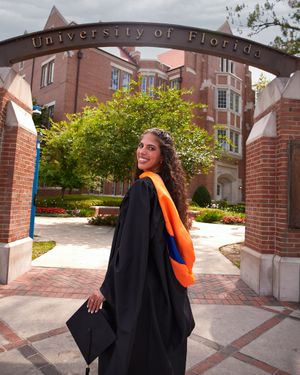 Graduate in black gown with orange stole poses beneath University of Florida arch.