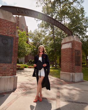 Graduate in black gown poses beneath University of Florida arch in elegant stance.