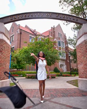 Graduate celebrates under University of Florida entrance arch in white dress.